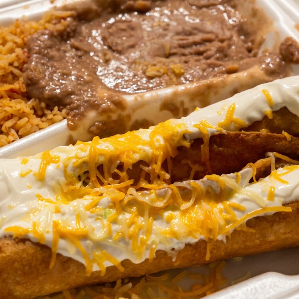 Taquitos topped with sour cream and shredded cheese, served with rice and refried beans in a takeout container.