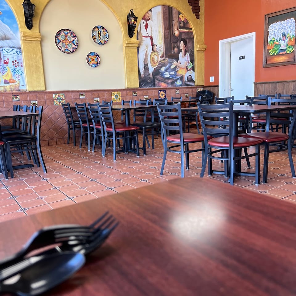 Vibrant restaurant dining room with Mexican murals, tiled floor, and empty tables with black and red chairs.