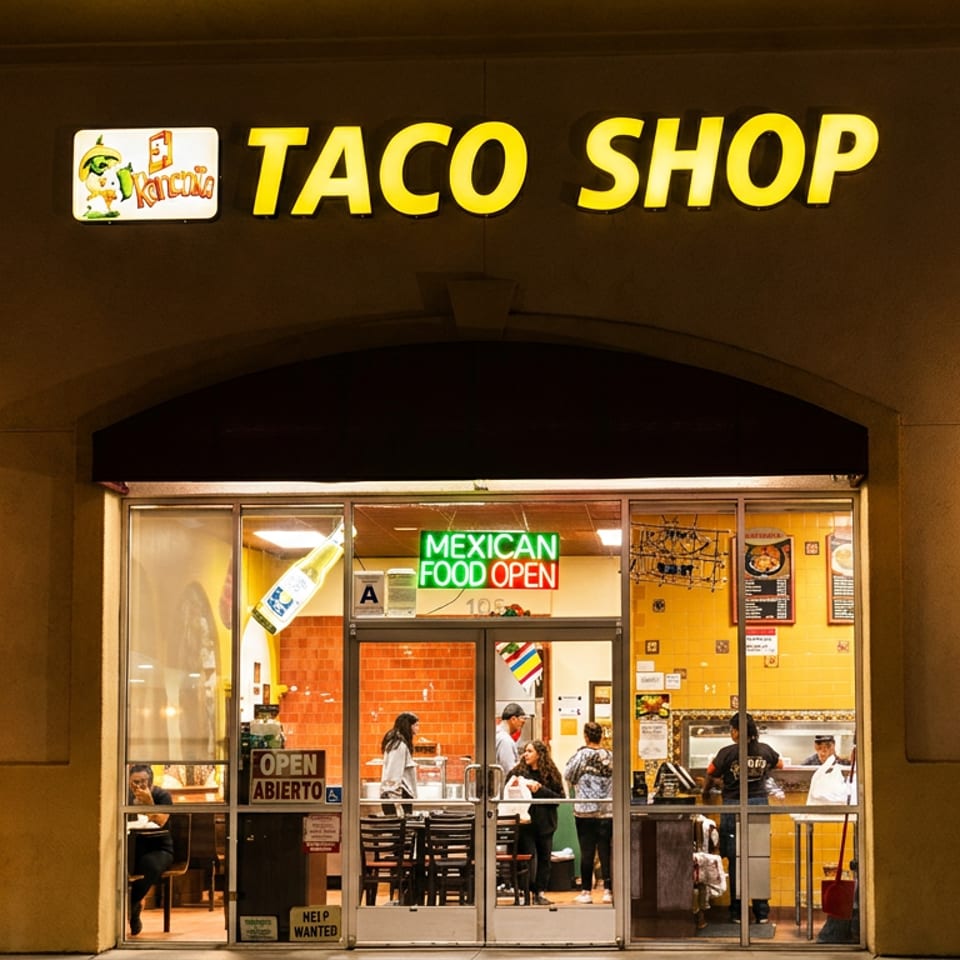 Nighttime exterior of a Taco Shop Mexican restaurant, with illuminated signage, open doors, and customers visible inside.